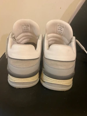 Axel Arigato sneakers i vitt och grått - Säljer ett par snygga Axel Arigato sneakers i vitt och ljusgrått med beige sula. Skorna har snörning, rund tå och detaljer i mocka och skinn. Märket syns på sidan och sulan. Perfekta för dig som gillar stilrena och trendiga sneakers.