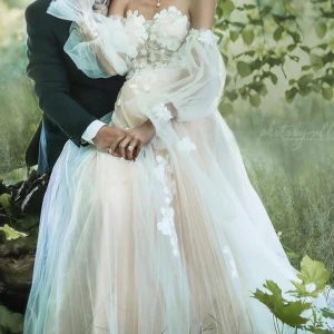 Champagnefärgad långklänning med blommor - Drömmig champagnefärgad långklänning med puffiga långärmar i tunn tyll och vackra vita broderade blommor över hela klänningen. Klänningen har snörning i ryggen och transparenta detaljer som ger en romantisk vibe. Perfekt för dig som vill sticka ut med en unik och elegant look.