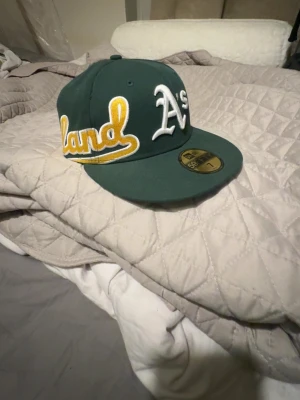 Grön Oakland Athletics keps - Säljer en grön 59FIFTY keps från New Era med Oakland Athletics broderat i vitt och gult. Klassisk rak skärm i grönt och detaljerad MLB-logga baktill. Kepsen är tillverkad i 100% polyester och har en snygg streetstil som passar perfekt till din outfit.