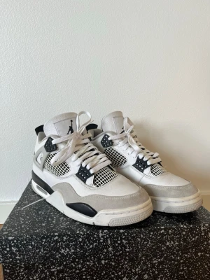 Nike Air Jordan 4 military black - Säljer ett par Nike Air Jordan 4 i vitt, svart och grått. Skorna har klassisk meshpanel, detaljer i mocka och skinn samt ikoniska Jumpman-loggan på plösen och hälen. Snygg snörning och robust sula med svart och vit kontrast. Perfekta för dig som vill sticka ut med en tidlös sneaker.