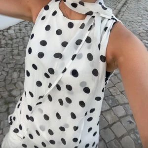 Vit prickig tröja med scarf - Säljer en vit tröja med svarta prickar och matchande scarf runt halsen. Den är ärmlös och har en luftig passform. Materialet är lätt och ger en skön känsla mot huden. Snygg och trendig look