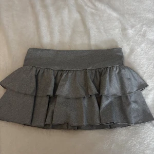 Grå volangkjol 🩶 - Supersöt grå kortkjol med dubbla volanger. Kjolen har en mjuk och stretchig känsla, perfekt för dig som gillar en lekfull och trendig stil. Passar dig som vill ha något enkelt men ändå med lite extra detaljer.