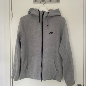 Nike tech fleece - Nike tech fleece med svart logga på bröstet. Tröjan har hel dragkedja, huva med snörning och två stora fickor framtill. Perfekt för chill eller träning, och tillverkad i mjuk bomullsblandning för extra komfort. Har en liten fläck på vänster ficka som borde gå bort i tvätten.