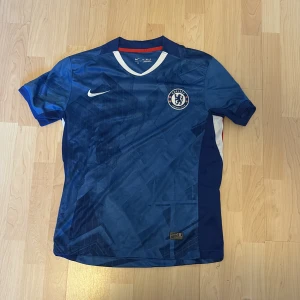 Chelsea FC blå Nike fotbollströja - Chelsea FC matchtröja från Nike i blått med diskret geometriskt mönster. Tröjan har klubbmärke på bröstet, vit V-ringning och röda samt vita detaljer på sidorna. Tillverkad i lätt och ventilerande material, perfekt för fotboll eller träning.