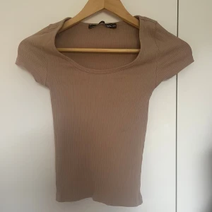 Beige ribbad topp med kort ärm - En enkel och stilren beige ribbad topp. Toppen har korta ärmar, rundad halsringning och är figurnära i modellen. Perfekt att matcha med jeans eller kjol för en clean look.