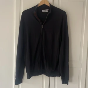 Svart kofta från Dressmann i bomull - Svart kofta från Dressmann i mjuk pima bomull. Tröjan har hel dragkedja framtill, ribbade muddar och klassisk passform. Perfekt för lager-på-lager och funkar året runt. Enkel och stilren design utan mönster.