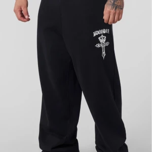 Svarta mjukisbyxor med tryck - Svarta mjukisbyxor med vitt tryck på låret som visar en korsdesign och texten 'YOUNGLA'. Byxorna har en loose passform och är tillverkade i mjukt material som passar chill och streetwear. Perfekt för dig som gillar avslappnad stil.