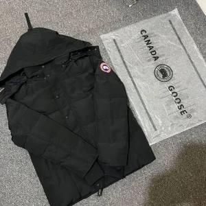 Svart dunjacka från Canada Goose - Säljer en svart dunjacka från Canada Goose med klassisk patch på ärmen. Jackan har huva, långa ärmar och stängs med både dragkedja och knappar. Perfekt för kalla dagar och riktigt snygg streetstil.