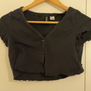 Svart croppad topp med v-ringning - Svart croppad topp med v-ringning och knapp framtill. Toppen har korta ärmar och råa kanter nertill för en trendig look. Perfekt att matcha med höga jeans eller kjol.