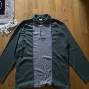 Nike pikétröja grå och mörkgrön - Nike pikétröja med lång ärm i grått och mörkgrönt. Klassisk krage, knäppning med fyra knappar och broderad Nike-logga på bröstet. Tröjan har en bred grå panel framtill och mörkgröna sidor och ärmar. Perfekt för en sportig och avslappnad stil.