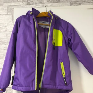 Lila vindjacka från Snowtex med huva - Säljer en lila vindjacka från Snowtex med huva och coola neongula detaljer på dragkedjor och bröstficka. Jackan har långa ärmar, praktiska fickor med dragkedja och är tillverkad i syntetmaterial. 