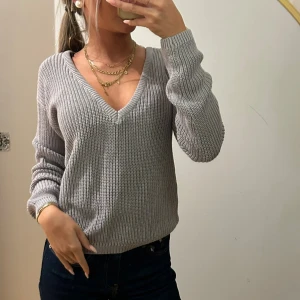 Grå stickad v-ringad tröja - Snygg grå stickad tröja med djup v-ringning och långa ärmar. Tröjan har en ribbad struktur och kan bäras både på axlarna eller offshoulder för en chill vibe. Perfekt att matcha med jeans och smycken för en trendig look.