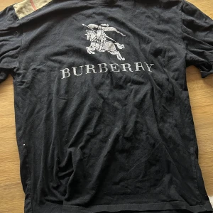 Svart Burberry t-shirt med logga - Svart t-shirt från Burberry med stor klassisk ryttare och Burberry-logga tryckt på bröstet. T-shirten har korta ärmar och rund halsringning, samt en detalj i Burberrys rutiga mönster på axeln. Tillverkad i mjuk bomull.