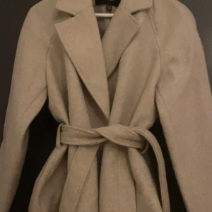 Beige kappa med bälte och krage - Stilren beige kappa med bred krage och knytskärp i midjan. Jackan har lång ärm och en klassisk, rak passform som ger en snygg siluett. Perfekt för dig som gillar en clean och tidlös look.