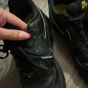 Nike Air Force 1 svart med gula detaljer - Svarta Nike Air Force 1 sneakers med snygga gula detaljer på loggan och swoosh. Låg modell med klassisk rund tå och perforerad ovandel i skinn. Svart platt sula med tydlig Nike-mönstring undertill. Skorna har snörning och gul insida. Stl 39 använt men fint skick