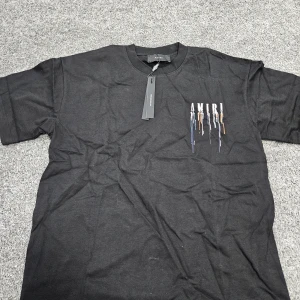 Svart AMIRI t-shirt med färgstänk - Svart t-shirt från AMIRI med coolt tryck av droppande färg i blått, orange, vitt och lila både fram och bak. Oversized fit och klassisk rund hals. Perfekt statement-plagg för dig som gillar streetwear och unika detaljer.
