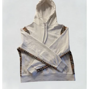 Burberry hoodie - Burberry hoodie – vit med klassiskt rutmönster  Exklusiv och tidlös hoodie från Burberry i vitt med de ikoniska rutiga detaljerna längs ärmarna och sidorna. Tillverkad i mjuk, högkvalitativ bomull som ger en lyxig känsla och perfekt passform.  Detaljer: 	•	Märke: Burberry 	•	Färg: Vit med beigea detaljer 	•	Material: 100 % bomull 	•	Passform: Regular fit 	•	Storlek: S/XS 	•	Skick: Mycket bra skick – inga slitningar, hål eller fläckar  Stilren, bekväm och lätt att matcha 