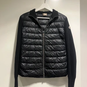 Svart quiltad cardigan Moncler - Svart cardigan från Moncler med quiltad front och rygg i glansigt material, stickade ärmar och krage. Jackan har dragkedja framtill och Moncler-logga på vänster ärm. Perfekt för dig som vill ha en clean och stilren look.