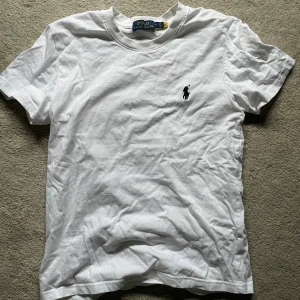 Vit t-shirt från Polo Ralph Lauren - Klassisk vit t-shirt från Polo Ralph Lauren med broderad svart logga på bröstet. T-shirten har rund halsringning och korta ärmar. Tillverkad i mjuk bomull för en skön och avslappnad passform.