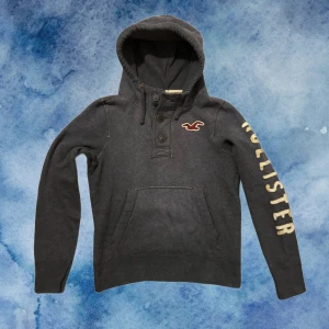Vintage Hollister Hoodie Med Knappar - Mysig mörkgrå hoodie från Hollister med broderad logga på bröstet och stor text längs ena ärmen. Tröjan har huva med snörning, knappar vid halsen och en stor magficka. (pyttelitet hål vid luvan men syns ej då hela hooduen har en sliten design) 