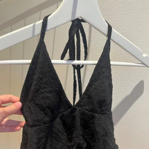 Svart volangig linnetopp med knyt - Säljer en svart linnetopp med volanger och knyt i nacken. Den är lite nopprig men annars inget fel.