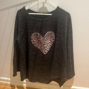 Mörkgrå tröja med leopardhjärta - Mysig mörkgrå långärmad tröja med ett stort hjärta i leopardmönster på bröstet. Tröjan har rund halsringning och är tillverkad i ett mjukt, stickat material som passar perfekt när du vill vara både chill och snygg.