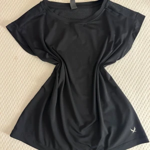 Svart träningstopp  - Svart träningstopp  med meshpaneler över axlarna och ryggen för extra ventilation. Toppen är ärmlös och har en loose passform, perfekt för gymmet eller löprundan. Materialet är lätt polyester och loggan syns diskret framtill.