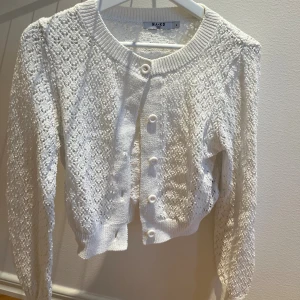 Vit stickad kofta från NA-KD - Superfin vit stickad kofta från NA-KD med genombrutet mönster och rund halsringning. Koftan har långa ärmar, ribbade muddar och knäppning framtill med stora vita knappar. Perfekt att slänga över en topp för en söt look.