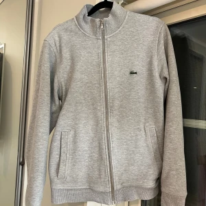 Grå ziptröja från Lacoste - Tja! Säljer min Lacoste ziptröja, då jag inte har någon användning utan av den längre.Snygg grå ziptröja från Lacoste med klassisk ståkrage och dragkedja hela vägen. Tröjan har två fickor fram och den ikoniska krokodilloggan broderad på bröstet. Perfekt för lager-på-lager och chill vardagsstil. Det är storlek S.