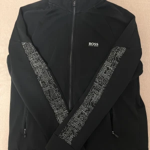  Hugo Boss tröja/kofta - Svart zipjacka från Hugo Boss med vita grafiska detaljer längs ärmarna och diskret logga på bröstet. Tröjan har hel dragkedja, hög krage och två fickor med dragkedja. Perfekt för en clean och modern streetstil. St : S , köpt för 80 euro pga det var en rabatt från Tyskland. 