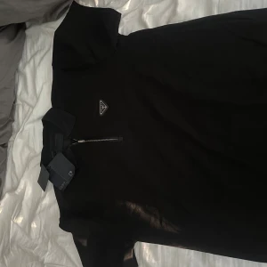 Svart pikétröja från Prada med zip - Svart pikétröja från Prada med klassisk krage och halv dragkedja framtill. Tröjan har korta ärmar och ikonisk Prada-logga på bröstet. Snygg och stilren design i bomull som passar perfekt till många olika looks.