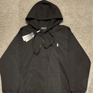 Ralph Lauren hoodie - Svart hoodie från Polo Ralph Lauren med klassisk huva och snörning. Tröjan har en vit broderad logga på bröstet och en stor magficka framtill. Tillverkad i mjuk bomull, perfekt för chill dagar. Stilren och enkel design som funkar till allt.