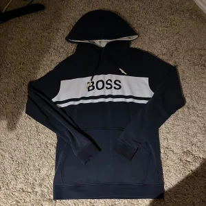 Mörkblå hoodie från BOSS med vit detalj - Snygg mörkblå hoodie från BOSS med vit panel och text över bröstet. Tröjan har huva med snörning, känguruficka och långa ärmar. Perfekt för en chill och stilren look.