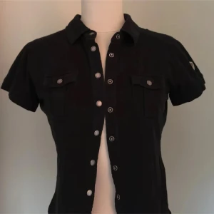 Svart kortärmad skjorta med knappar - Svart skjorta med korta ärmar och snygga silverfärgade knappar hela vägen framtill. Två bröstfickor med lock och knappdetaljer ger en cool touch. Skjortan har klassisk krage och är figurnära i modellen, perfekt för dig som gillar stilren och enkel design.