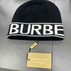 Svart stickad Burberry mössa - Svart stickad mössa från Burberry med vit, bred uppvikt kant och stor logotyp i vitt. Klassisk och stilren design som ger en cool vibe. Materialet är mjukt och värmande, perfekt för kyliga dagar. London England-taggen sitter kvar.