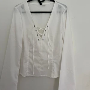 Vit långärmad blus med snörning - Säljer SNYGGASTE vita långärmade blusen med djup v-ringning och snygg snörningsdetalj framtill. Toppen har en figurnära passform och trumpetärmar. Ger en lite korsettliknande look verkligen skitsnygg. Aldrig använd utan endast testad och slutsåld!!!