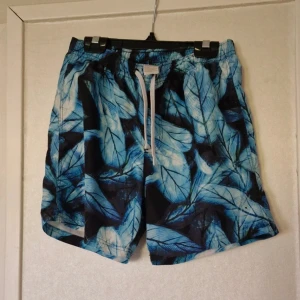 Blå mönstrade badshorts med snörning - Säljer badshorts med blått och svart bladmönster. De har vit snörning i midjan och är tillverkade i lätt polyester som torkar snabbt. Perfekta för stranden eller poolen, riktigt fräsch design som sticker ut.