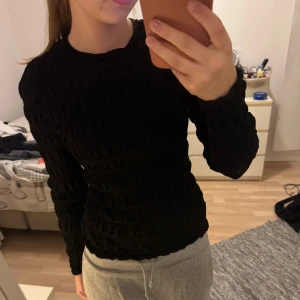 Svart ribbad långärmad tröja - Säljer en svart långärmad tröja med ribbad struktur och tight passform. Tröjan har rund hals och är tillverkad i ett mjukt syntetmaterial som sitter snyggt mot kroppen. Perfekt för dig som gillar stilrena och enkla plagg med lite extra detalj.