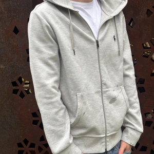 Ralph Lauren zip hoodie - Snygg ljusgrå hoodie från Ralph Lauren med dragkedja och klassisk svart logga på bröstet. Tröjan har huva med snörning, fickor framtill och är tillverkad i mjuk bomullsmix. Perfekt för chill dagar eller när du vill ha en clean look.