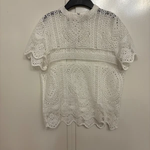 Vit spetsblus från Gina Tricot - Superfin vit blus från Gina Tricot med detaljerad spets och broderat mönster. Blusen har korta ärmar, hög hals och dragkedja bak. 