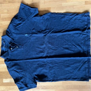 Mörkblå piké från Polo Ralph Lauren - Säljer en mörkblå pikétröja från Polo Ralph Lauren i bomull. Tröjan har klassisk krage, två knappar och den ikoniska broderade loggan på bröstet. Modellen är custom slim fit och passar perfekt för en clean och stilren look.