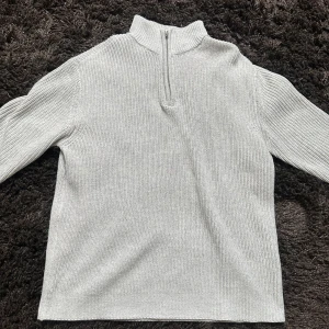 Ljusgrå stickad tröja med half zip - En stilren ljusgrå stickad tröja med hög krage och half zip-dragkedja framtill. Tröjan har långärmad passform och är ribbstickad i både ärmar och kropp. Perfekt för lager-på-lager under kyliga dagar.