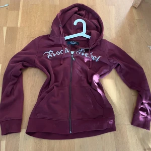 Vinröd hoodie Rock Rebel med tryck - Säljer en vinröd hoodie från Rock Rebel med dragkedja och huva. Framsidan har texten 'Rock Rebel' med glittriga detaljer och baksidan har ett stort tryck med kors och vingar. Mjuk insida och kängurufickor framtill. 