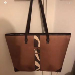 Brun shoppingväska med zebramönster - Stor brun shoppingväska i skinnimitation med långa mörkbruna axelband. Väskan har ett coolt zebramönstrat band i beige och brunt mitt på framsidan, samt en liten krona som detalj. Perfekt för dig som vill ha en rymlig och trendig väska med unik look.