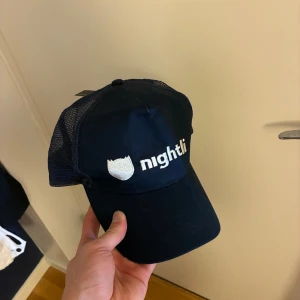 Nightli svart meshkeps med brodyr - Svart keps från Nightli med vit broderad logga framtill. Klassisk trucker-modell med böjd skärm och mesh på sidorna och bak för extra ventilation. Justerbar passform med plastspänne. Perfekt för dig som vill ha en clean och stilren look.