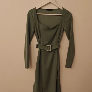 Boohoo klänning  - Snygg olivgrön klänning från Boohoo med lång ärm och markerad midja tack vare ett matchande bälte med silverfärgat spänne. Klänningen har en smickrande fyrkantig ringning och figurnära passform. Perfekt för dig som gillar stilrena och trendiga plagg.