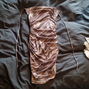 Brun mönstrad kortklänning med snörning - Säljer en kort tight klänning i bruna och beige toner med ett unikt randigt mönster. Klänningen har snörning i halsen och är figurnära med snygga rynkade detaljer längs hela längden. Materialet är mjukt och stretchigt, troligen polyester. Perfekt för dig som gillar trendiga och coola plagg.