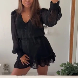 Svart kortklänning med volang - Supersnygg svart kortklänning med transparenta långärmar och prickigt mönster. Klänningen har v-ringning, knappar framtill och volangdetaljer vid nederkanten. Perfekt för dig som gillar en feminin och trendig look.