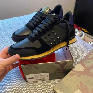 Valentino Garavani Rockrunner sneakers - Snygga svarta Valentino Garavani Rockrunner sneakers med coola nitar på sidorna och kontrasterande gul sula. Skorna har en mix av skinn och mocka, samt svarta snören och detaljer i lila vid hälen. Perfekt för dig som vill sticka ut med lyxiga detaljer.