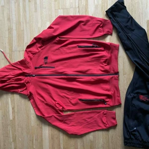 Röd hoodie från The North Face - Säljer en röd hoodie från The North Face med svarta detaljer och dragkedjor. Tröjan har huva med snörning, långa ärmar och flera praktiska fickor med dragkedja. Perfekt för dig som gillar sportig och avslappnad stil.